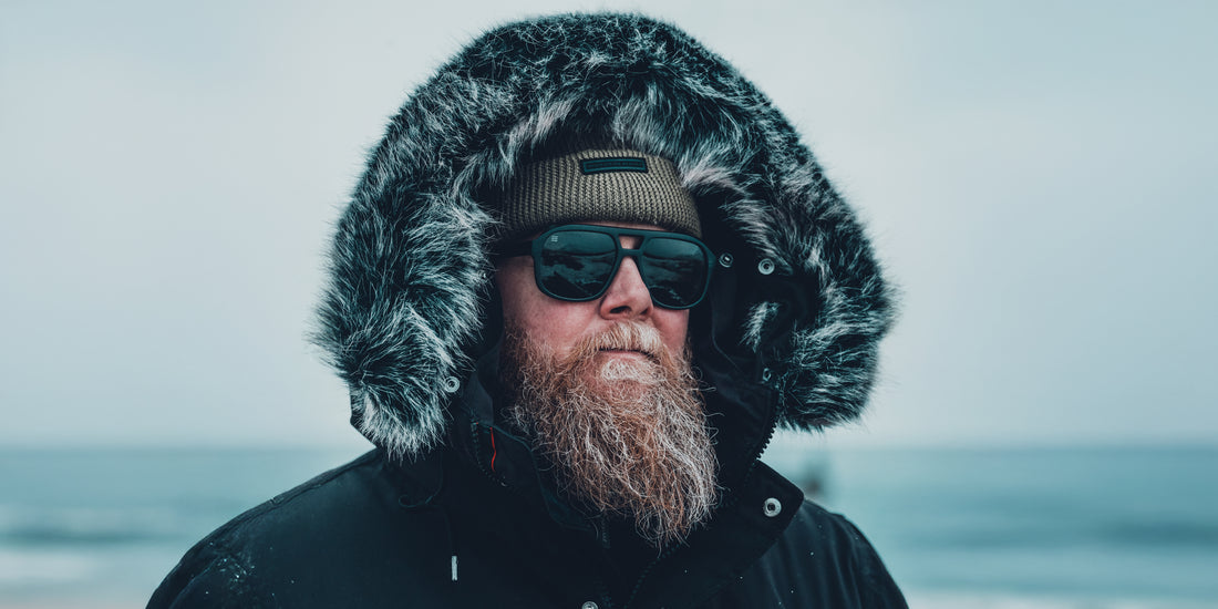 Bearded man wearing black Cosmo aviator polarized sunglasses and a hooded fur-trim parka on a cold seashore