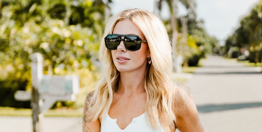 Blonde woman wearing black Cosmo aviator polarized sunglasses on a sunlit suburban street with palm trees in the background