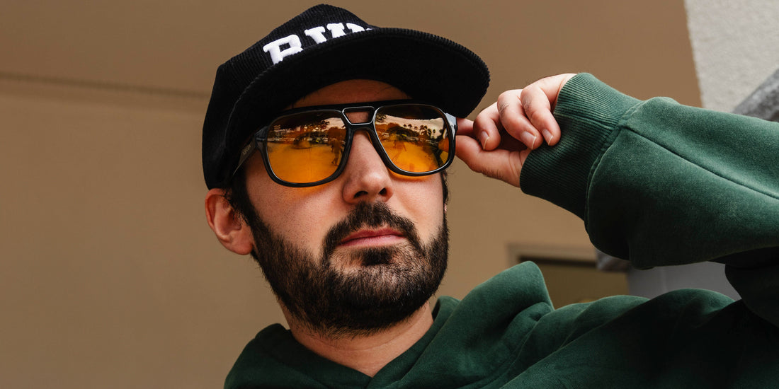 Man adjusting Helios black aviator sunglasses with yellow lenses while wearing a black cap and green sweatshirt