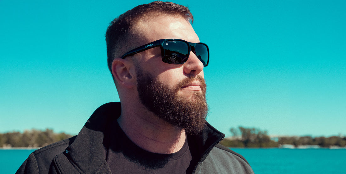Bearded man wearing Doom XL oversized glossy black sunglasses with polarized black lenses against a bright blue sky