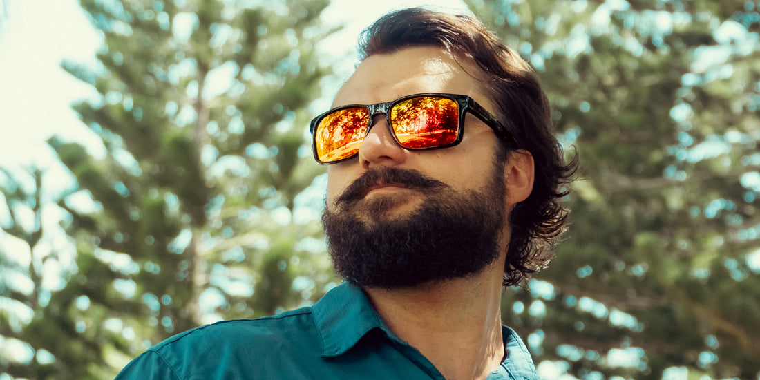 Man wearing John Wayne XL sunglasses, close up showing glossy black frame and red mirrored polarized lenses reflecting trees