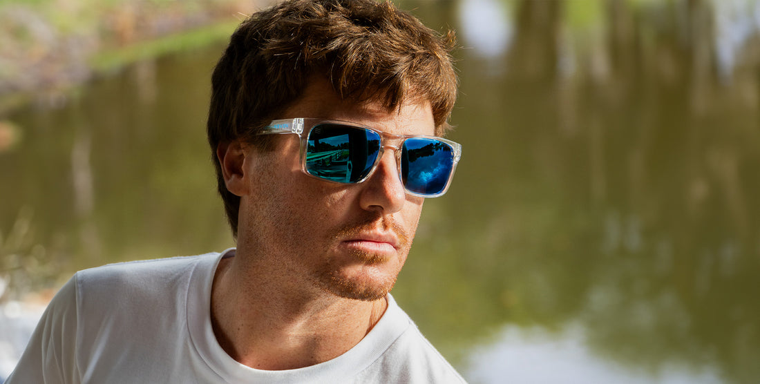 Man wearing Surge XL clear-frame sunglasses with blue mirrored polarized lenses, close-up portrait by a pond