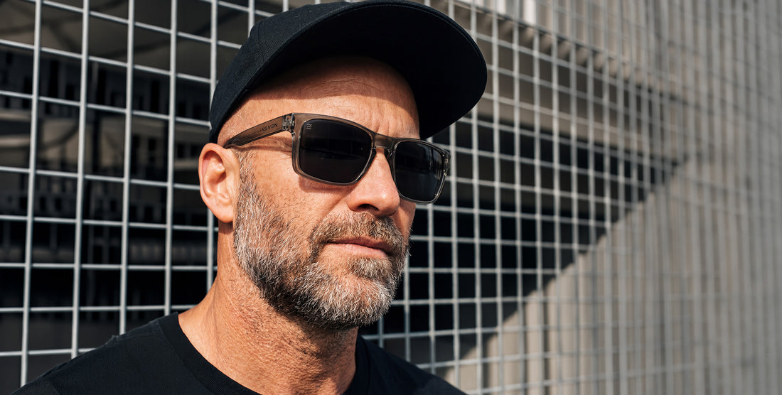 Three-quarter close-up of Breakers sunglasses with transparent gray glossy frame and black polarized lenses against a metal grid wall