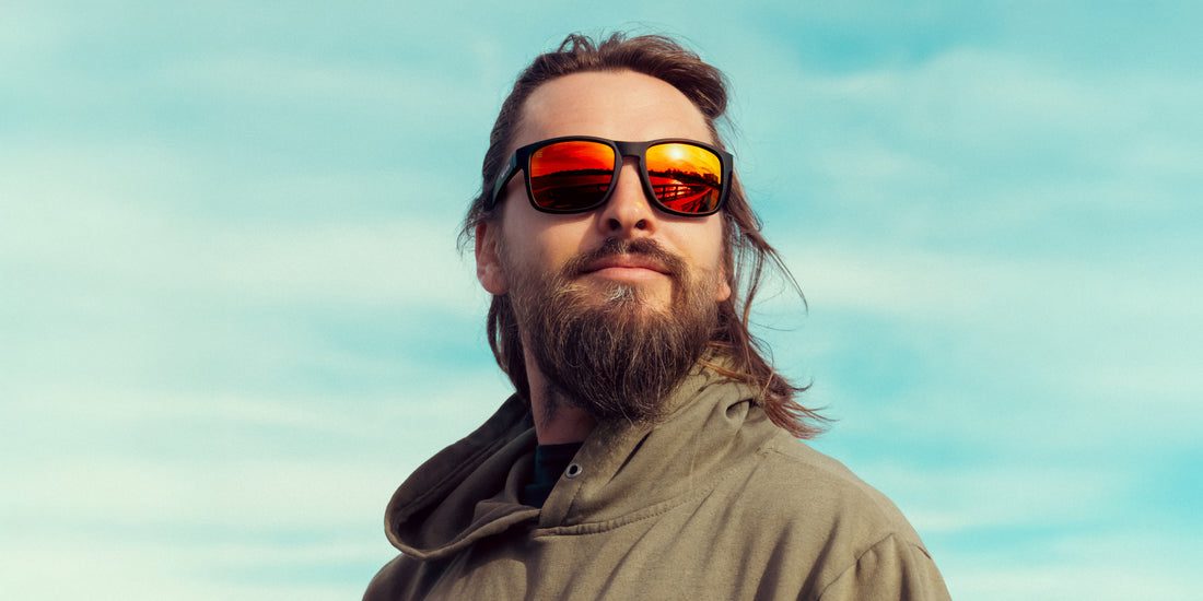 Bearded man wearing John Wayne black sunglasses with red mirrored lenses looking up at a blue sky