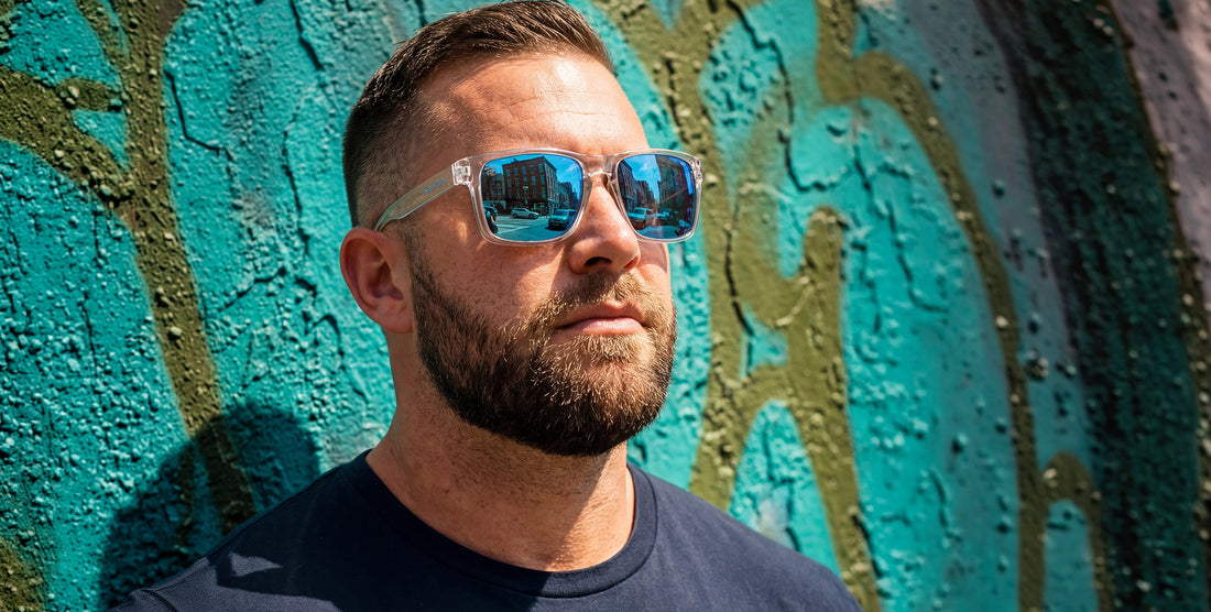 Man modeling Surge clear rectangular sunglasses with blue mirrored polarized lenses against a textured teal wall
