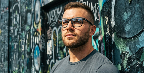 Man wearing matte black Agent Smith rectangular glasses looking up beside a graffiti-covered wall