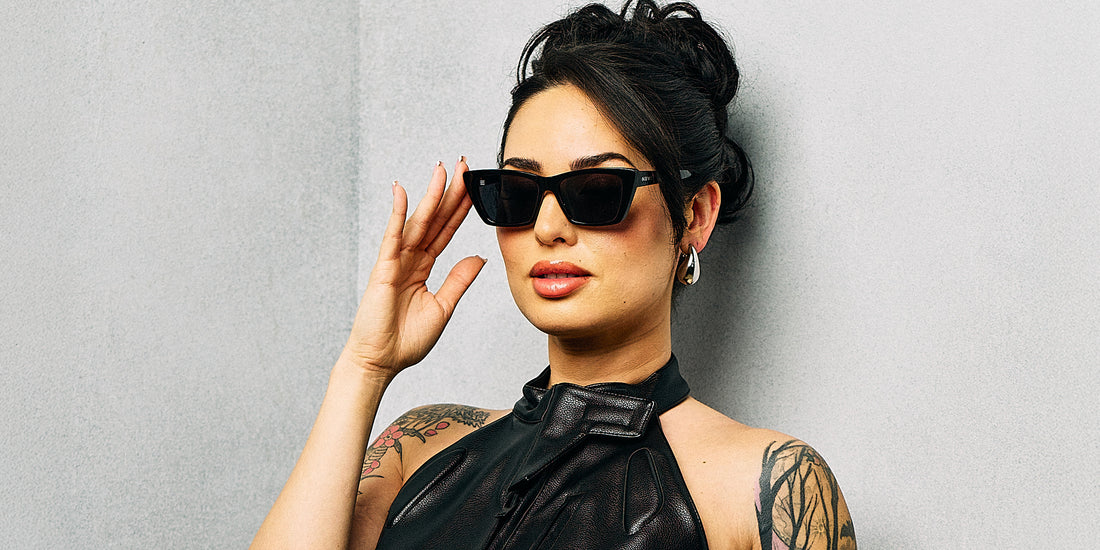 Model wearing Noir black cat-eye polarized sunglasses with silver temple accents, touching the frame against a light gray wall