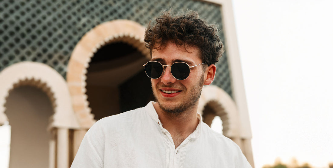 Man smiling wearing Rae gold round sunglasses with black lenses in front of a decorative arched wall