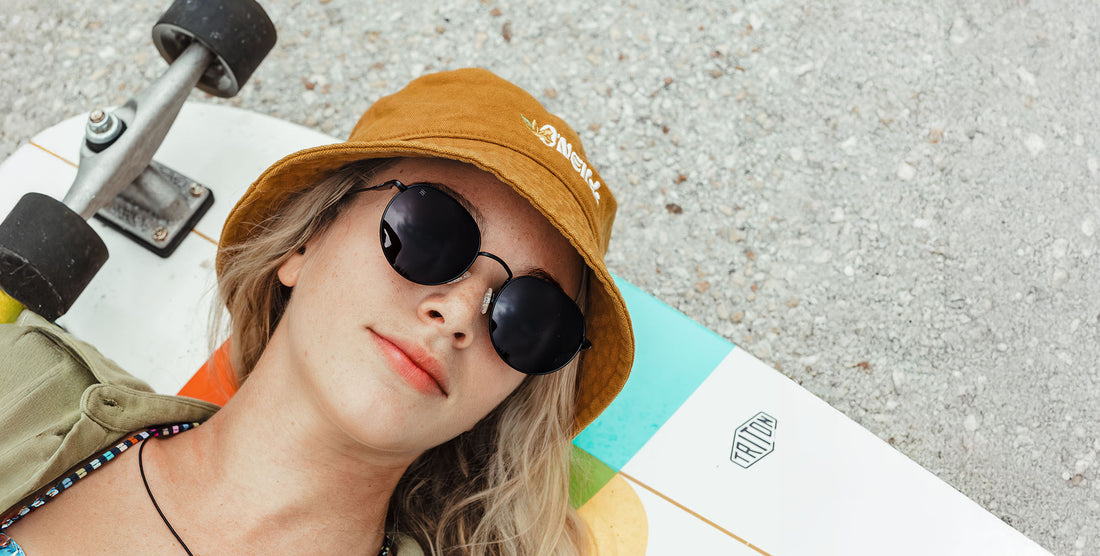 Woman wearing Saint black round polarized sunglasses and a mustard bucket hat, lying on a skateboard on gravel