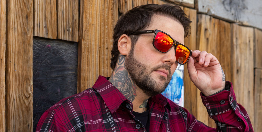 Man adjusting black Cinder sunglasses with red mirrored lenses outdoors by a rustic wooden wall
