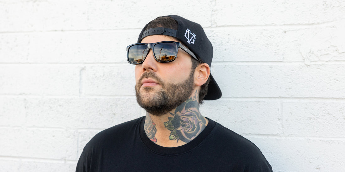 Three-quarter view of Triumph sunglasses showcasing large gloss black rectangular frame, polarized lenses with etched Phoenix logo and subtle silver accents on a model against a white brick wall