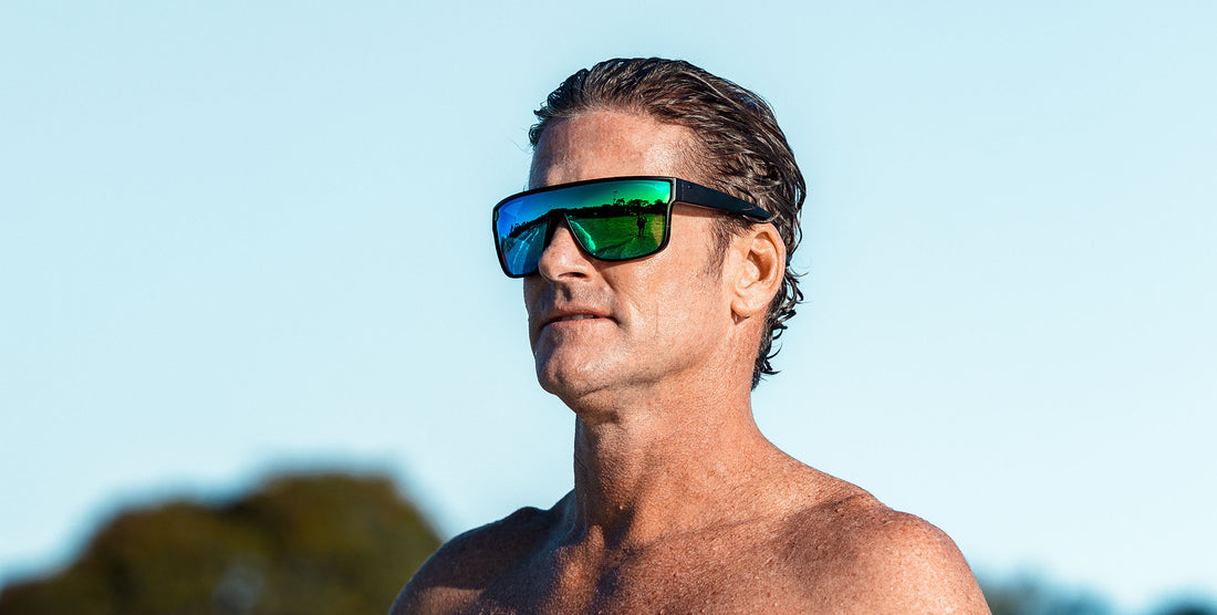 Three-quarter view of Valley sunglasses showcasing black matte frame and blue-green mirrored polarized shield lens on a male model against a clear blue sky