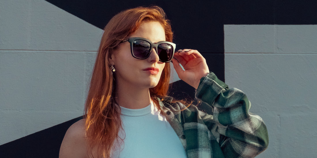 Woman adjusting matte black Lido cat-eye polarized sunglasses with purple gradient lenses against a black-and-white mural