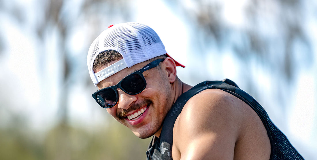 Smiling man wearing Classic black wayfarer polarized sunglasses and a white mesh cap outdoors with blurred palm trees behind him