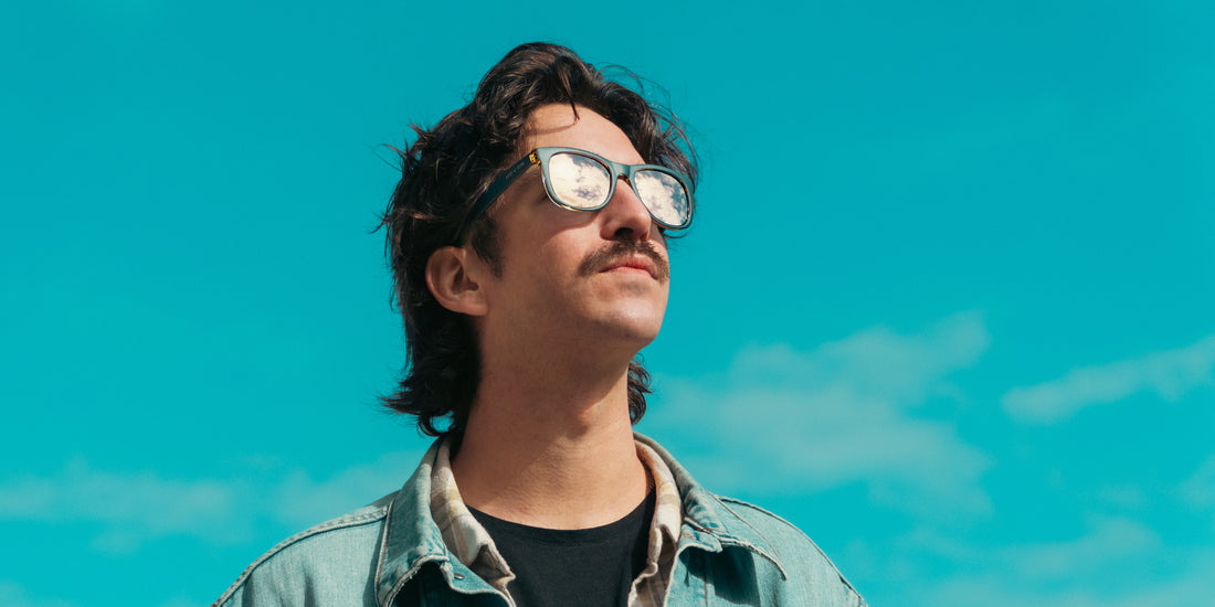 Man wearing Sundown blue rectangular polarized sunglasses looking up at a turquoise sky, clouds reflected in lenses