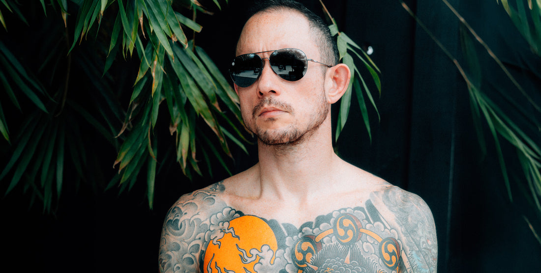 Tattooed man wearing Burakkumetaru aviator sunglasses against a dark backdrop with green palm leaves