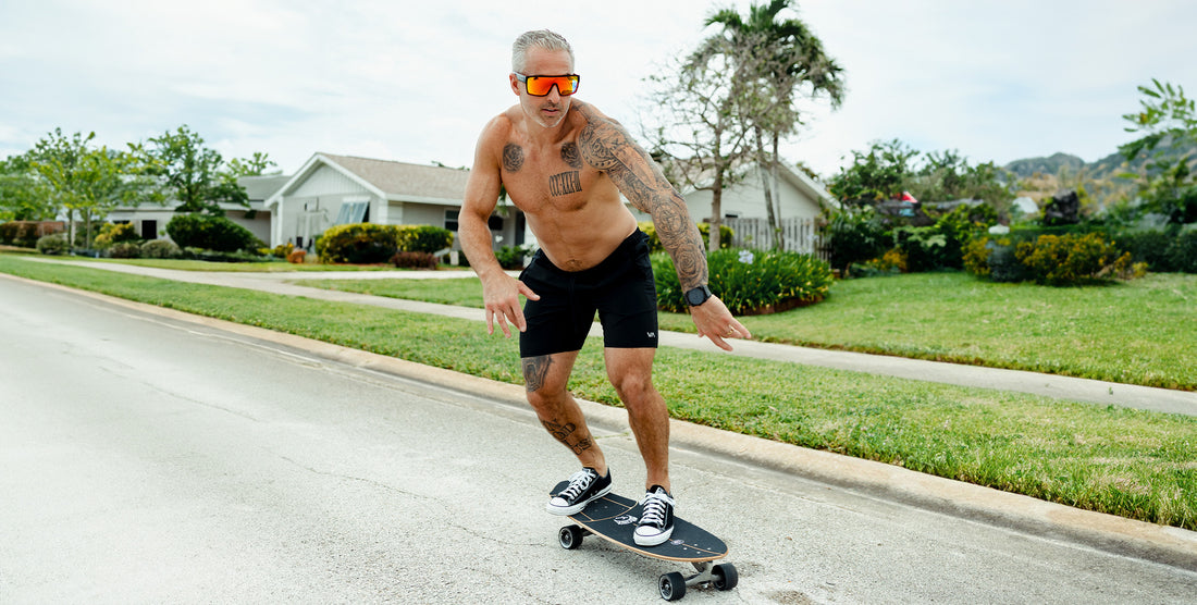Shirtless tattooed man skateboarding on a suburban street wearing 2nd Light black frame wraparound sunglasses with red-orange polarized lenses