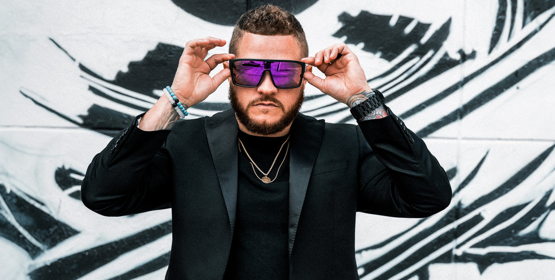 Man putting on Paradise polarized sunglasses with matte black frame and purple lenses in front of a black-and-white mural