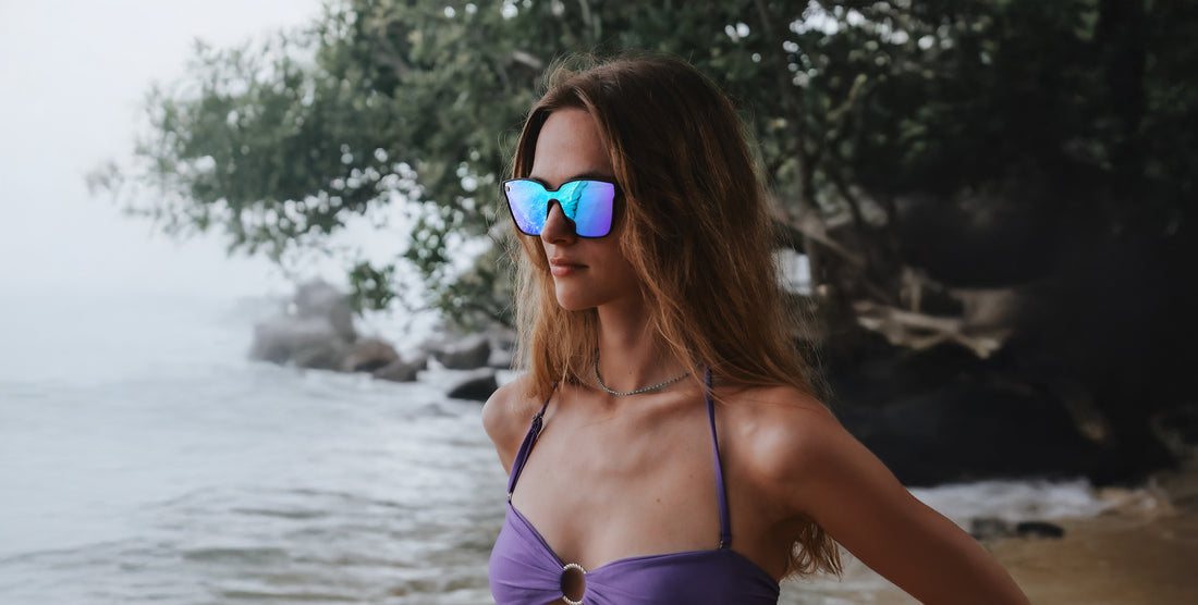Woman wearing Curiosity black cat-eye polarized sunglasses with blue-purple mirrored lenses standing on a misty beach