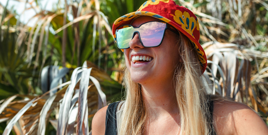 Woman smiling while wearing mirrored Endeavour sunglasses and a red floral bucket hat with sunlit palm leaves in the background
