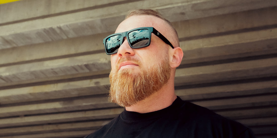 Man wearing Lowrider XL matte black polarized sunglasses looking up beneath a concrete overpass, lenses reflecting sky and palm trees