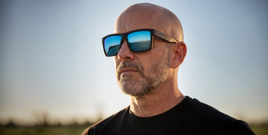 Angled close-up of Montauk XL sunglasses with matte black frame, mirrored blue polarized lenses and coffin logo, blurred background