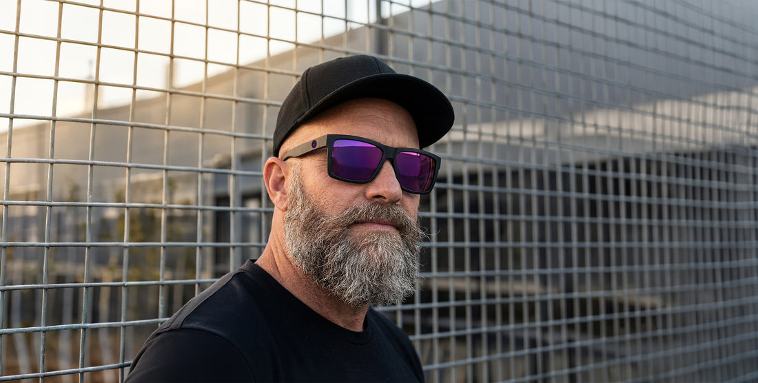 Bearded man wearing Nightshade XL matte black sunglasses with purple polarized lenses against a metal grid fence at sunset