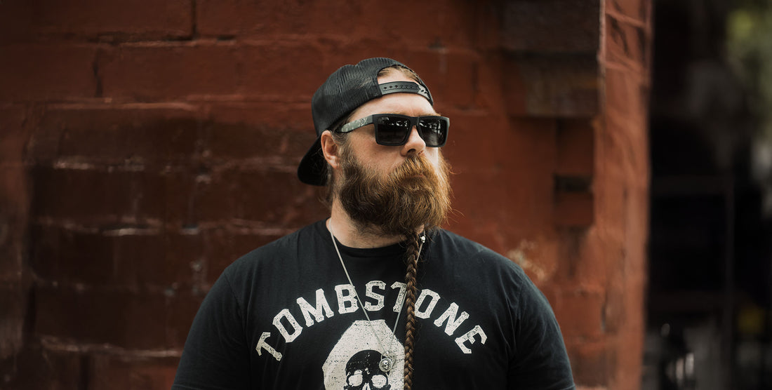 Bearded man wearing Bad Company polarized black sunglasses with camo-pattern arms, standing against a red brick wall