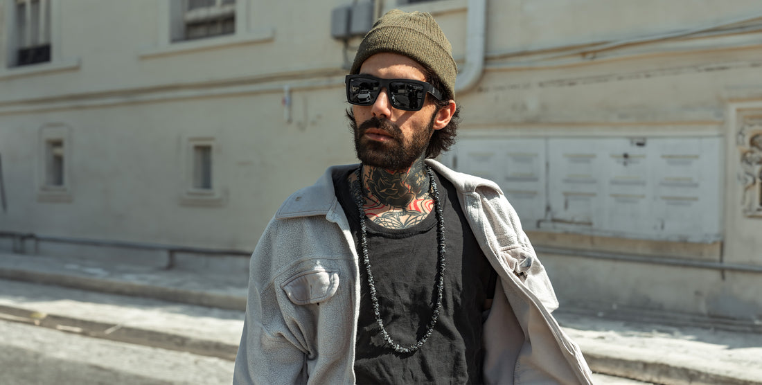 Bearded tattooed man wearing Lowrider black rectangular polarized sunglasses and an olive beanie on a sunlit urban street
