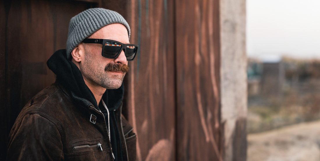 Bearded man wearing black Lowrider polarized sunglasses and a gray beanie beside a wooden building outdoors