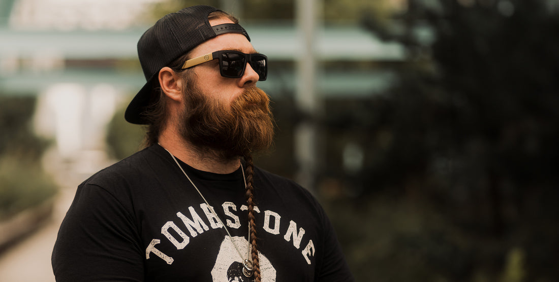 Bearded man wearing Prospector black polarized sunglasses with gold temple accents and backward mesh cap, looking right against a blurred outdoor background