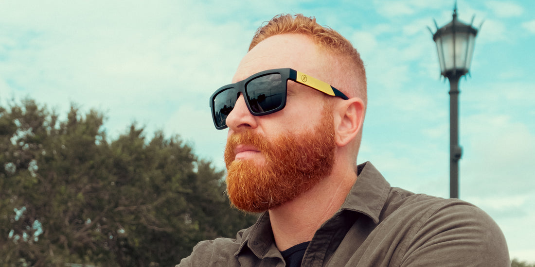 Man wearing Prospector XL oversized polarized sunglasses with black lenses and gold temples, looking into the distance outdoors with trees and a lamp post against a cloudy sky