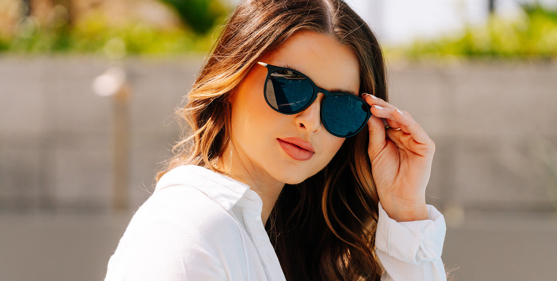 Woman wearing Goldie Lux black round polarized sunglasses adjusting them outdoors against a sunlit concrete wall