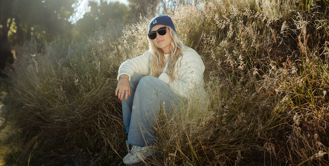 Woman sitting in sunlit grass wearing Goldie Lux black round polarized sunglasses and a blue beanie