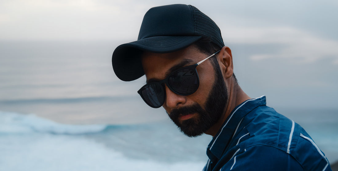 Man wearing Goldie Lux black round polarized sunglasses and a black cap looking over his shoulder with ocean waves in the background