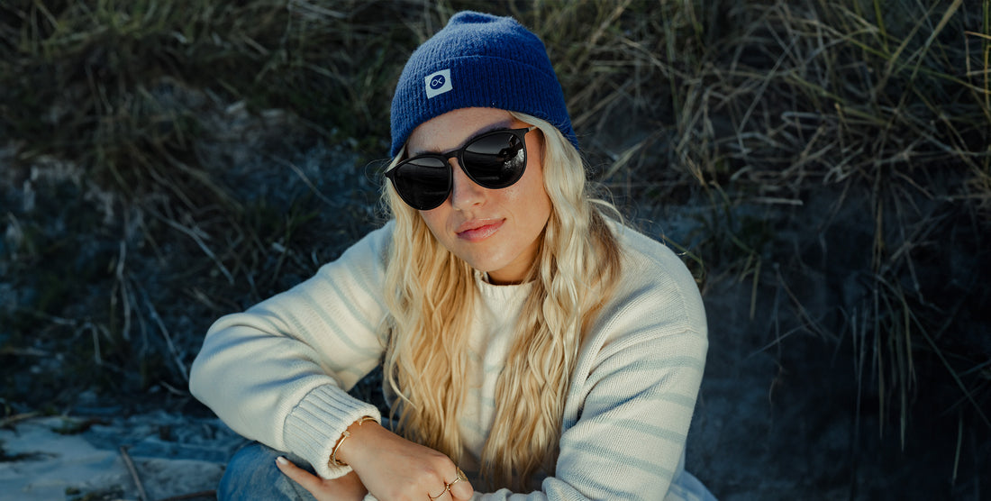 Woman wearing Goldie Lux black round polarized sunglasses and a blue beanie, seated on coastal rocks with tall grasses